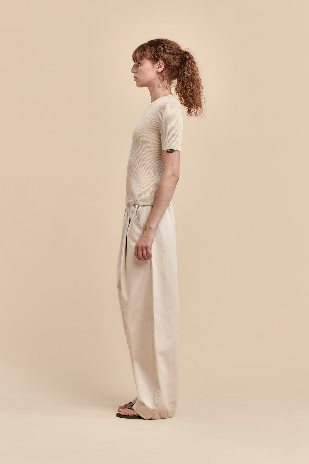 A model in profile wearing a beige short-sleeve knit top and matching wide-leg cotton trousers, standing against a solid beige studio background.
