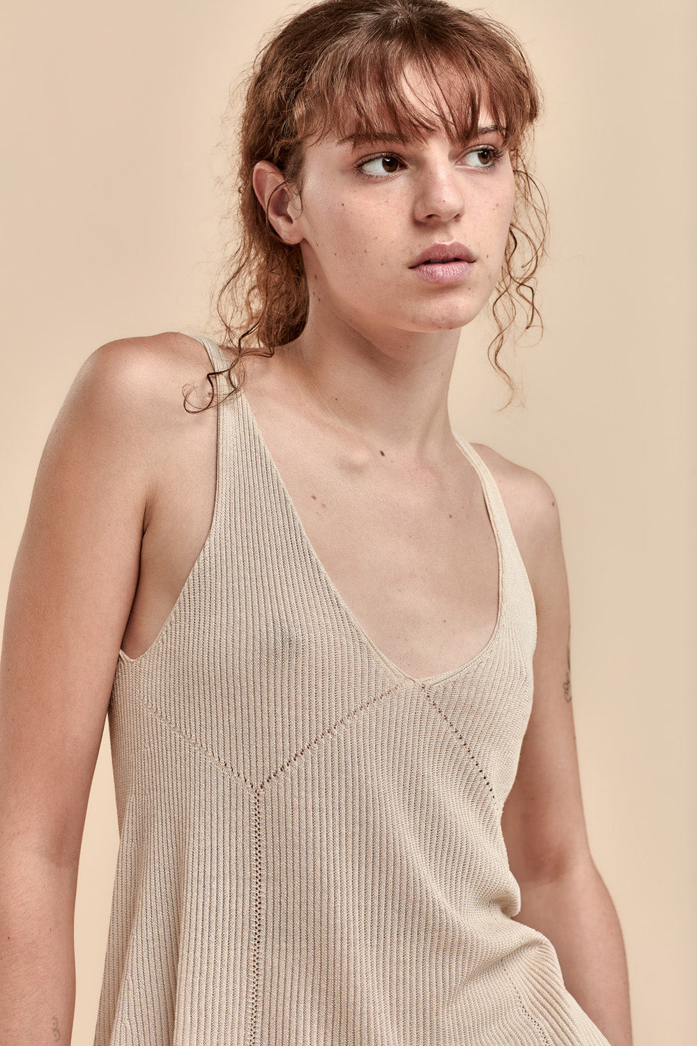 A woman wearing a beige ribbed knit camisole top with a deep V-neck and thin straps, photographed against a solid warm beige background.