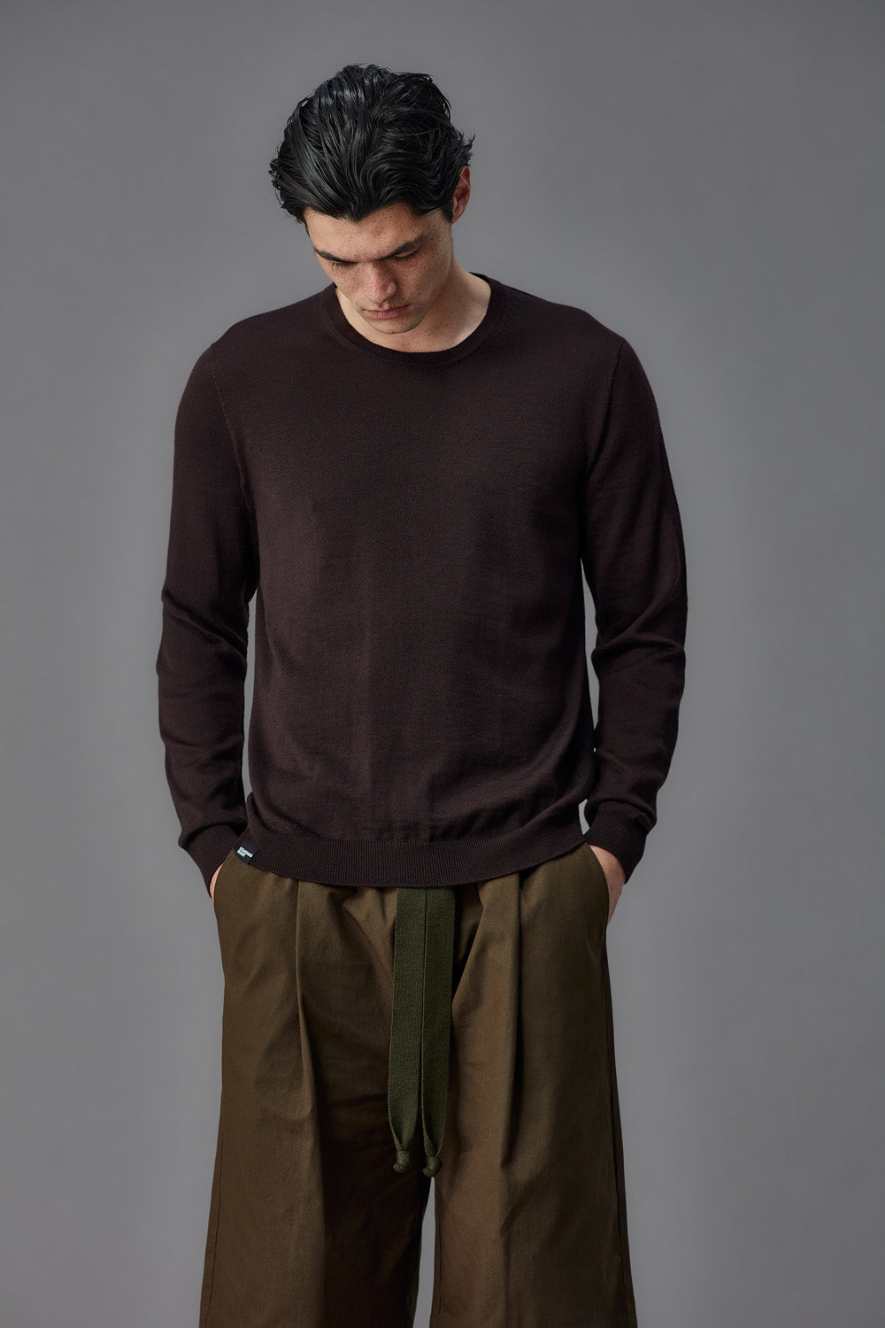 Man wearing a dark brown sweater and light brown pants against a gray background