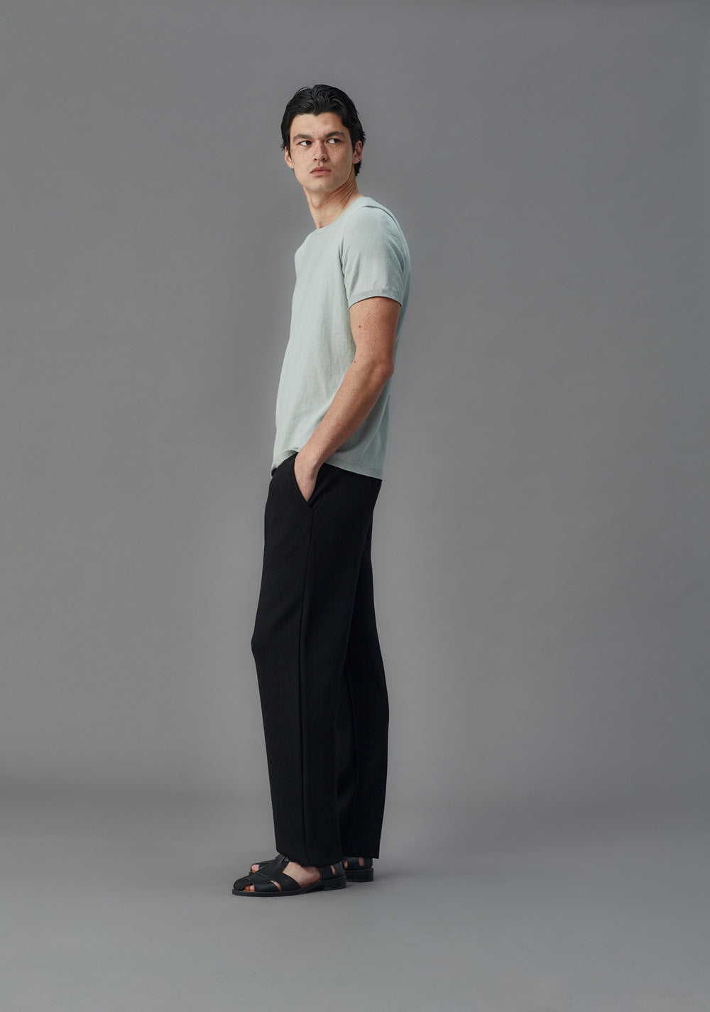 Man wearing black merino pants and a light blue t-shirt standing against a gray background