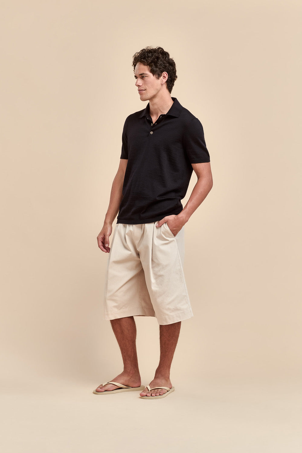 A man wearing a black merino short-sleeve polo shirt, off-white shorts, and beige flip-flops, standing against a solid beige background.
