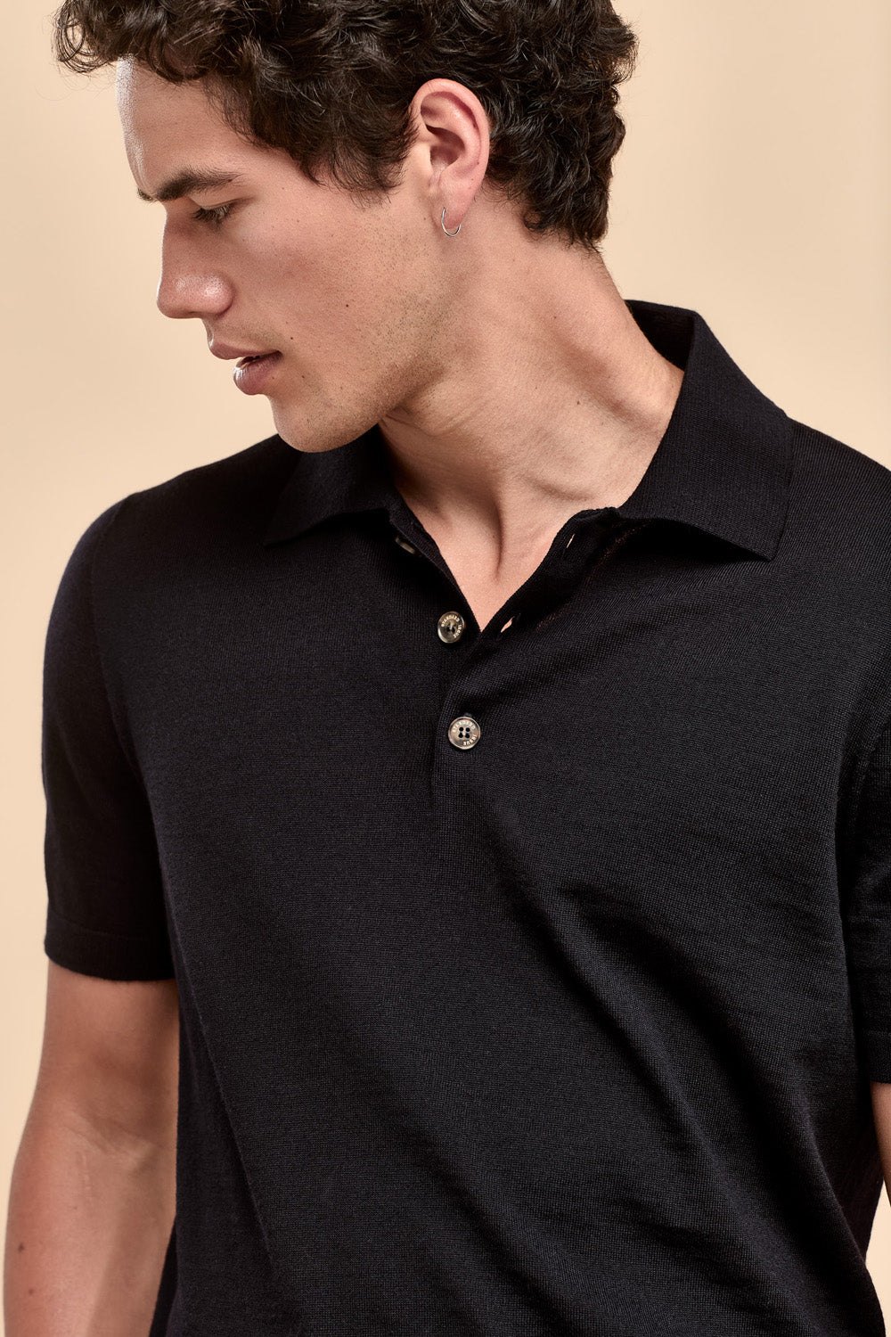 A close-up of a man wearing a black merino short-sleeve polo shirt with a three-buttons, set against a solid beige background.