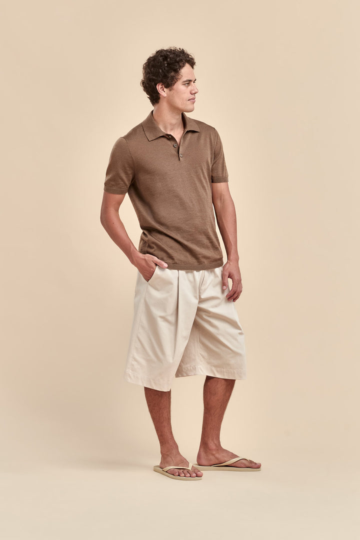 A man wearing a brown merino short-sleeve polo shirt and off-white shorts, standing in profile against a solid beige background.
