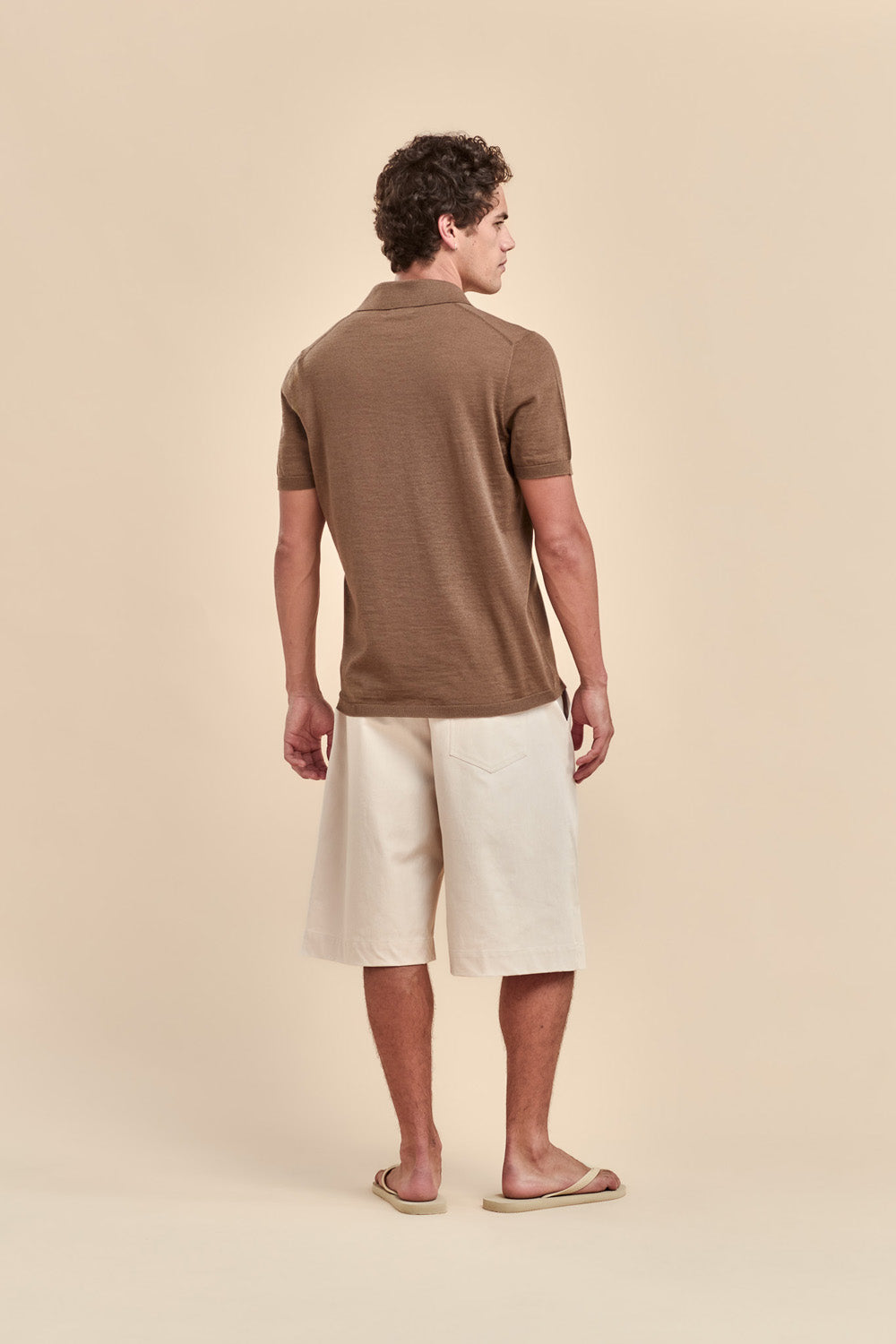 A man wearing a brown merino short-sleeve polo shirt and off-white shorts, standing in profile against a solid beige background.
