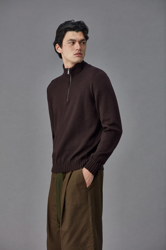 Man wearing a brown zip collar sweater and green pants against a gray background