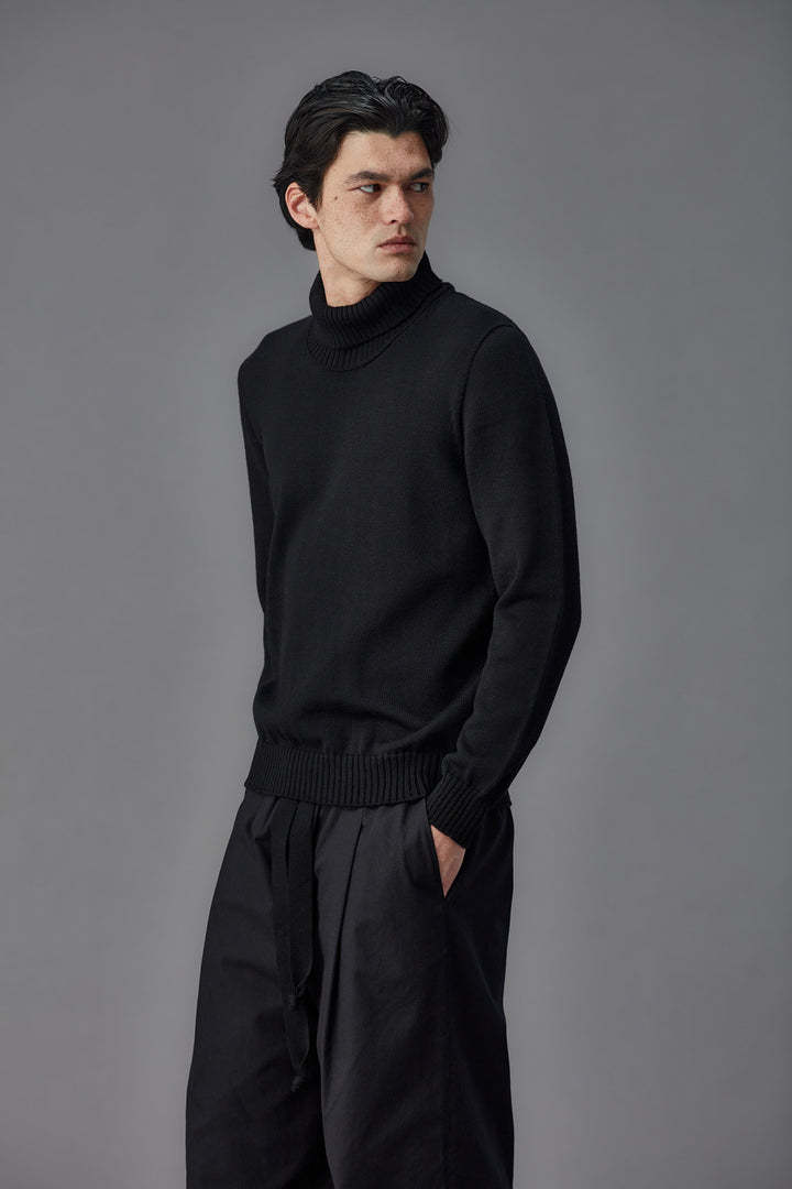 Man wearing a black turtleneck sweater and pants against a gray background