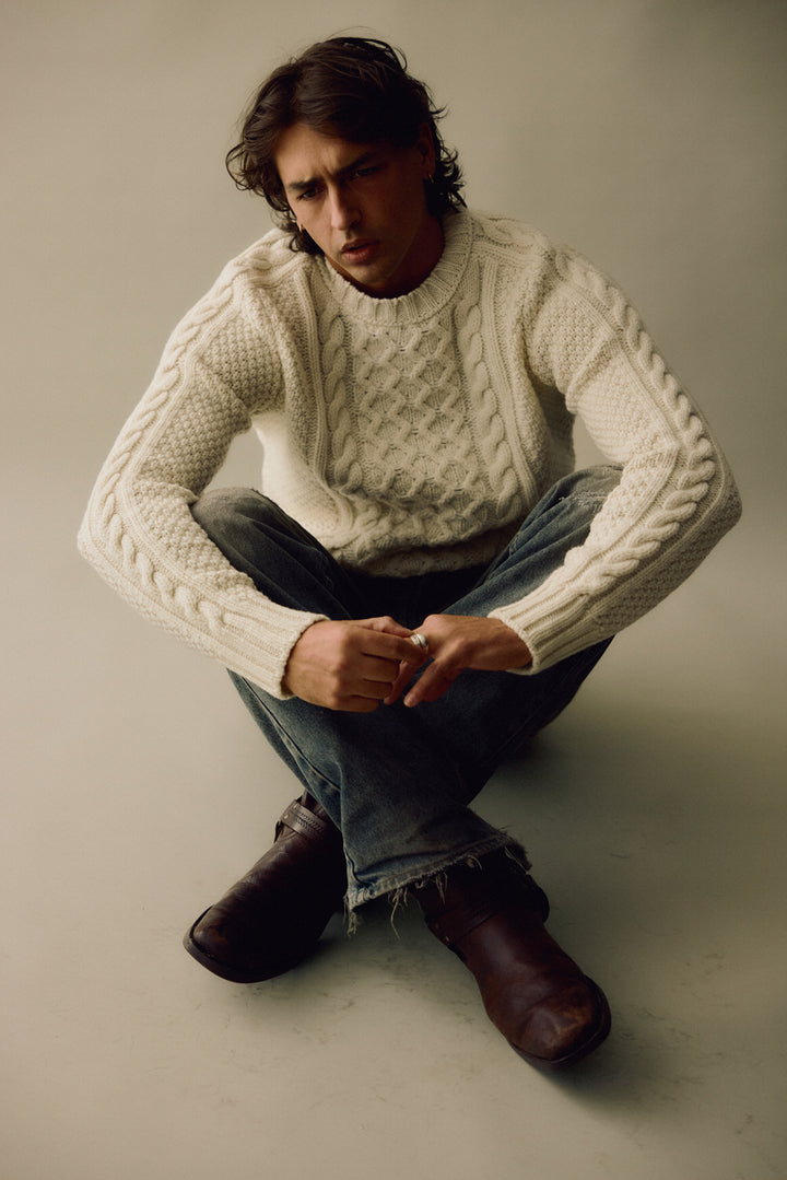Man wearing a white cable knit sweater and jeans sitting on the floor against a plain background